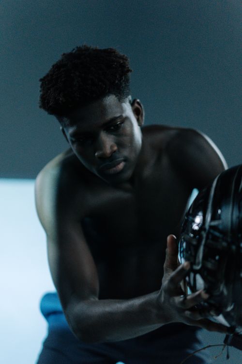 Free Shirtless young man gazing at a black helmet in a dimly lit indoor setting. Stock Photo