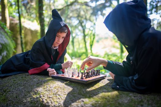 Two kids in wizard costumes play chess on a rock in a forest.