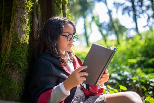 Free A young girl dressed in a costume reading a book while sitting by a mossy tree trunk in a forest. Stock Photo