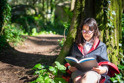 A Woman in Black Long Sleeves Dress Wearing Eyeglasses Sitting Under a Tree while Reading a Book