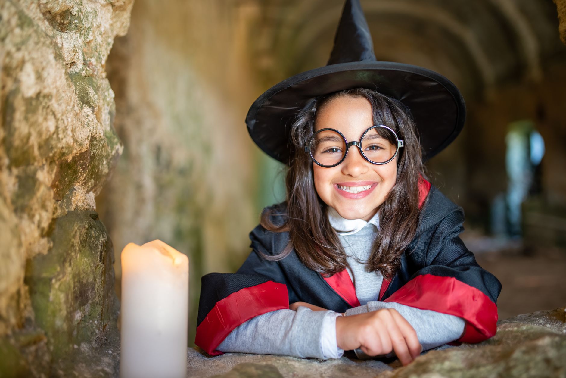 Smiling girl in witch costume with candle, set in ancient stone hallway, evokes magical charm.