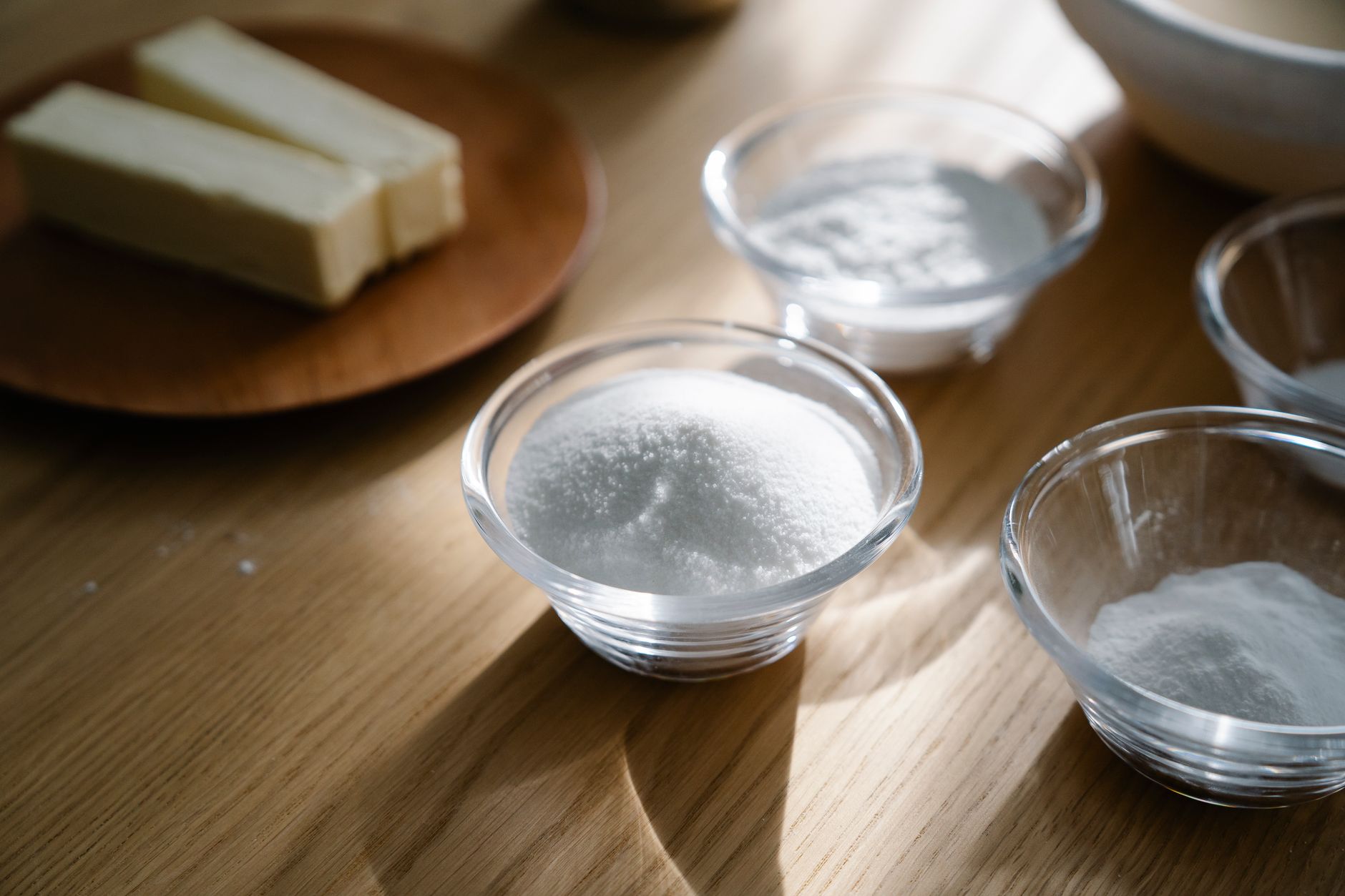 Glass bowls with sugar, flour, and butter on a wooden surface illuminated by natural light.