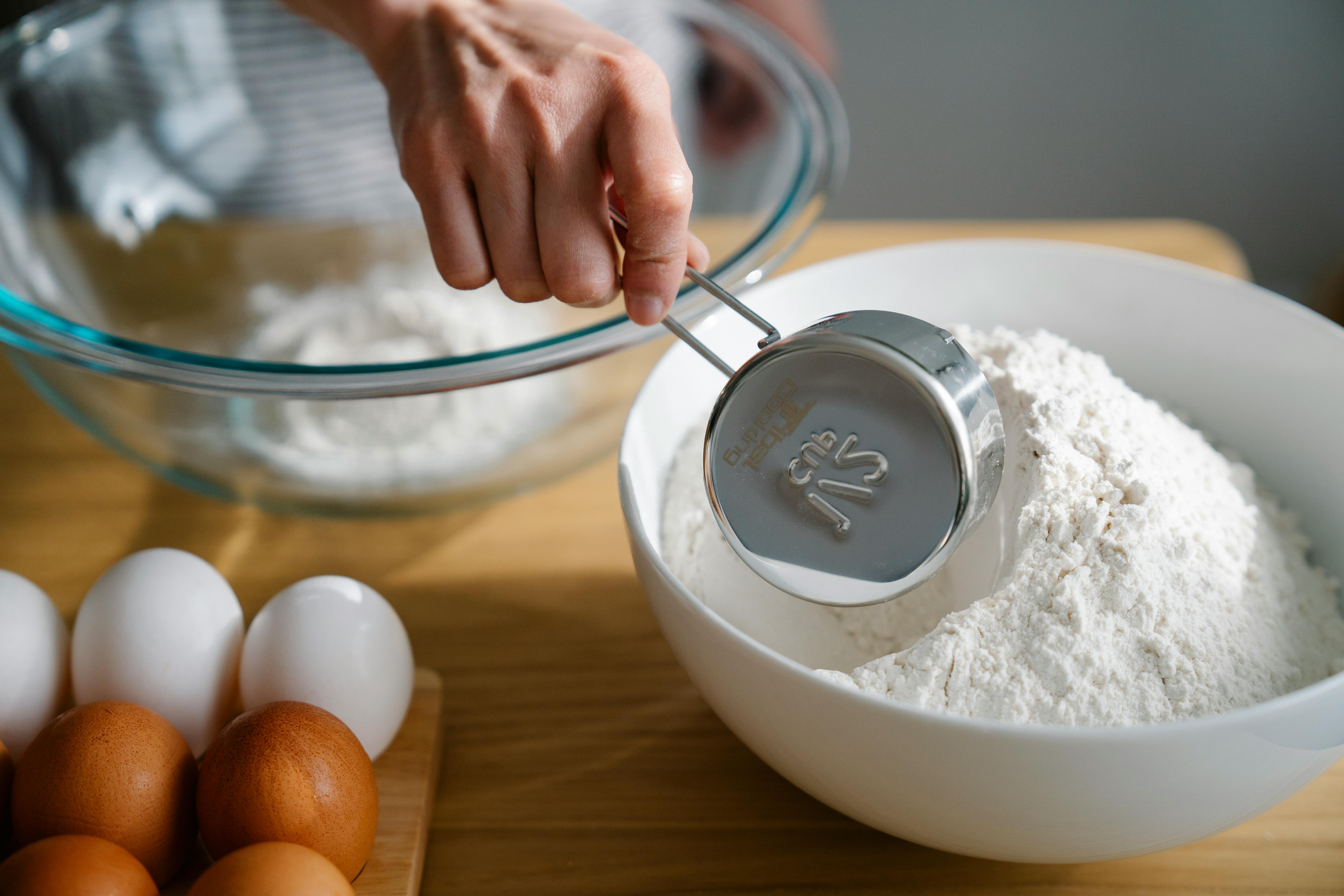 Close-up of a hand measuring flour in a bowl, ready for baking with eggs nearby on the kitchen counter.