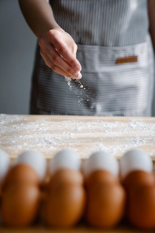 Free Close-up of a chef's hand sprinkling flour on a wooden surface with eggs nearby, preparing for baking. Stock Photo