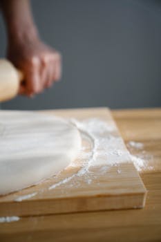 A hand rolling dough on a wooden board with flour, showcasing food preparation.