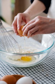 Close-up of hands cracking an egg into a bowl with a whisk, preparing ingredients for baking.