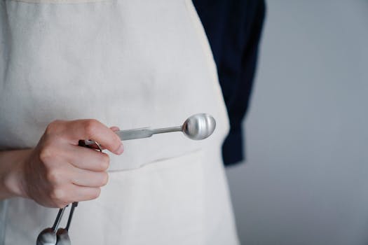 Close-up view of a baker's hand holding a measuring spoon in a kitchen setting.