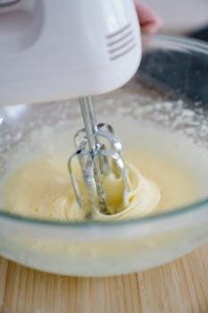 Close-up of an electric mixer blending creamy batter in a glass mixing bowl on a wooden surface.