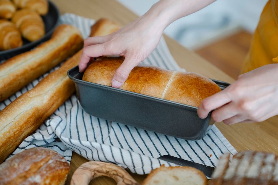 Hands holding a freshly baked loaf of bread in a pan, surrounded by various breads.