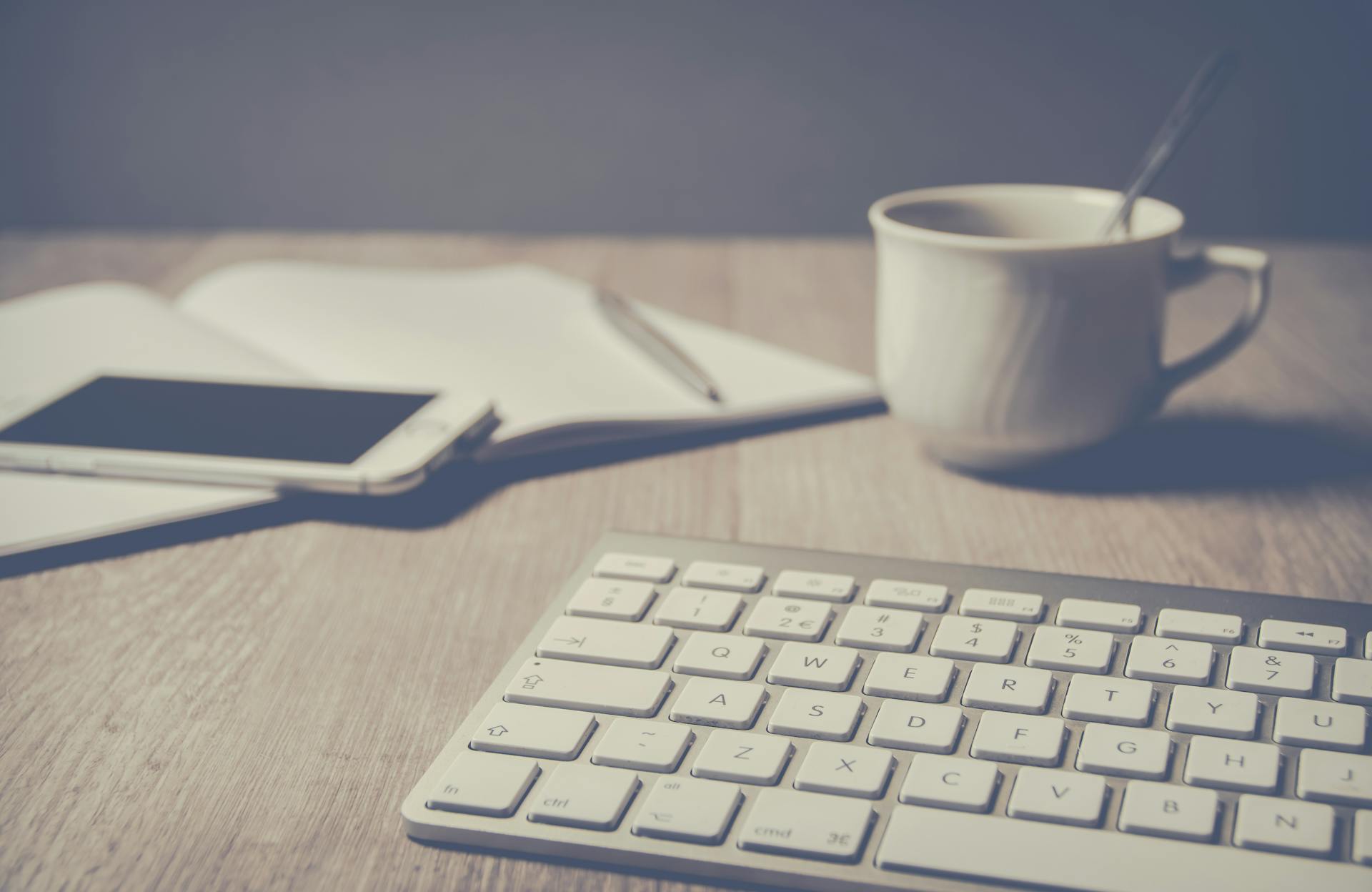 Cozy home office setup with keyboard, smartphone, notebook, and coffee cup on a wooden desk.