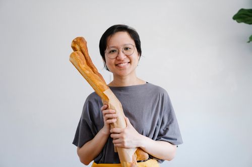 Free A cheerful woman in a gray shirt is smiling while holding a freshly baked baguette indoors. Stock Photo