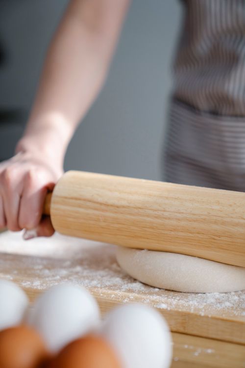 Free A baker uses a wooden rolling pin to flatten dough on a floured wooden board, with eggs nearby. Stock Photo