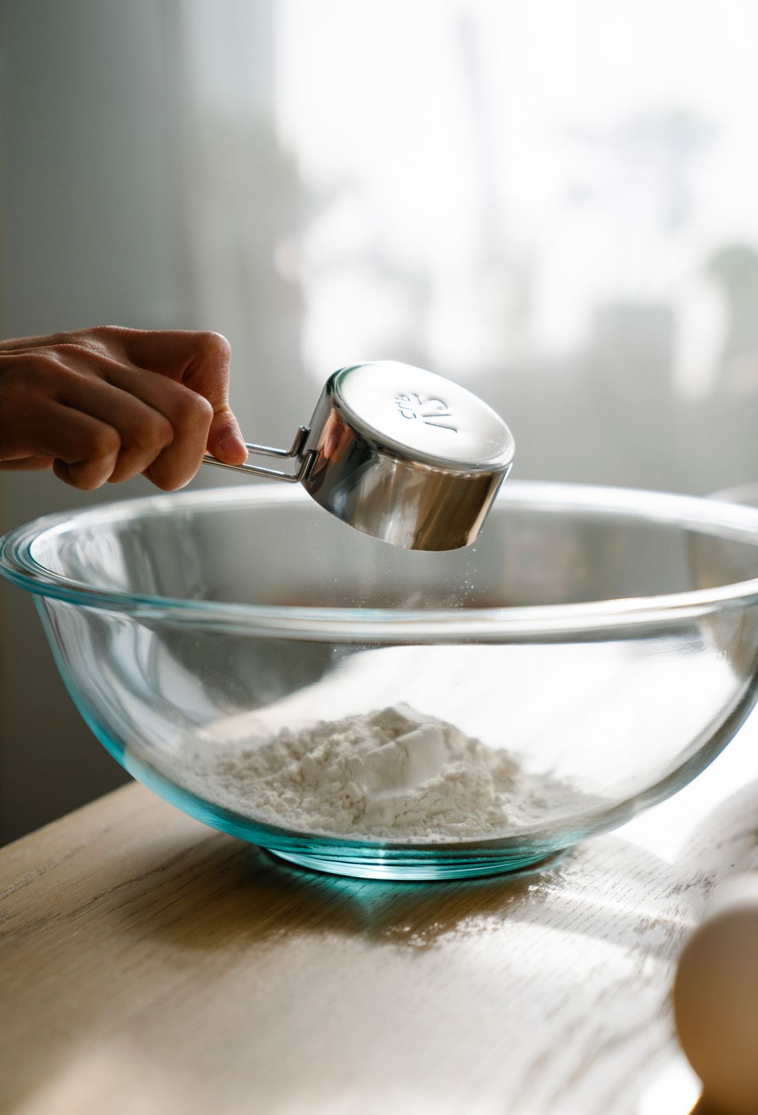 Clear glass mixing bowl with flour as a steel measuring cup pours