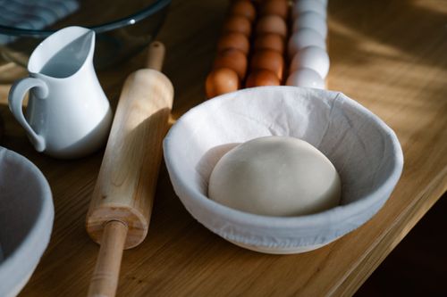 Free A bowl with dough near a rolling pin, jug, and eggs on a wooden table - perfect for baking themes. Stock Photo