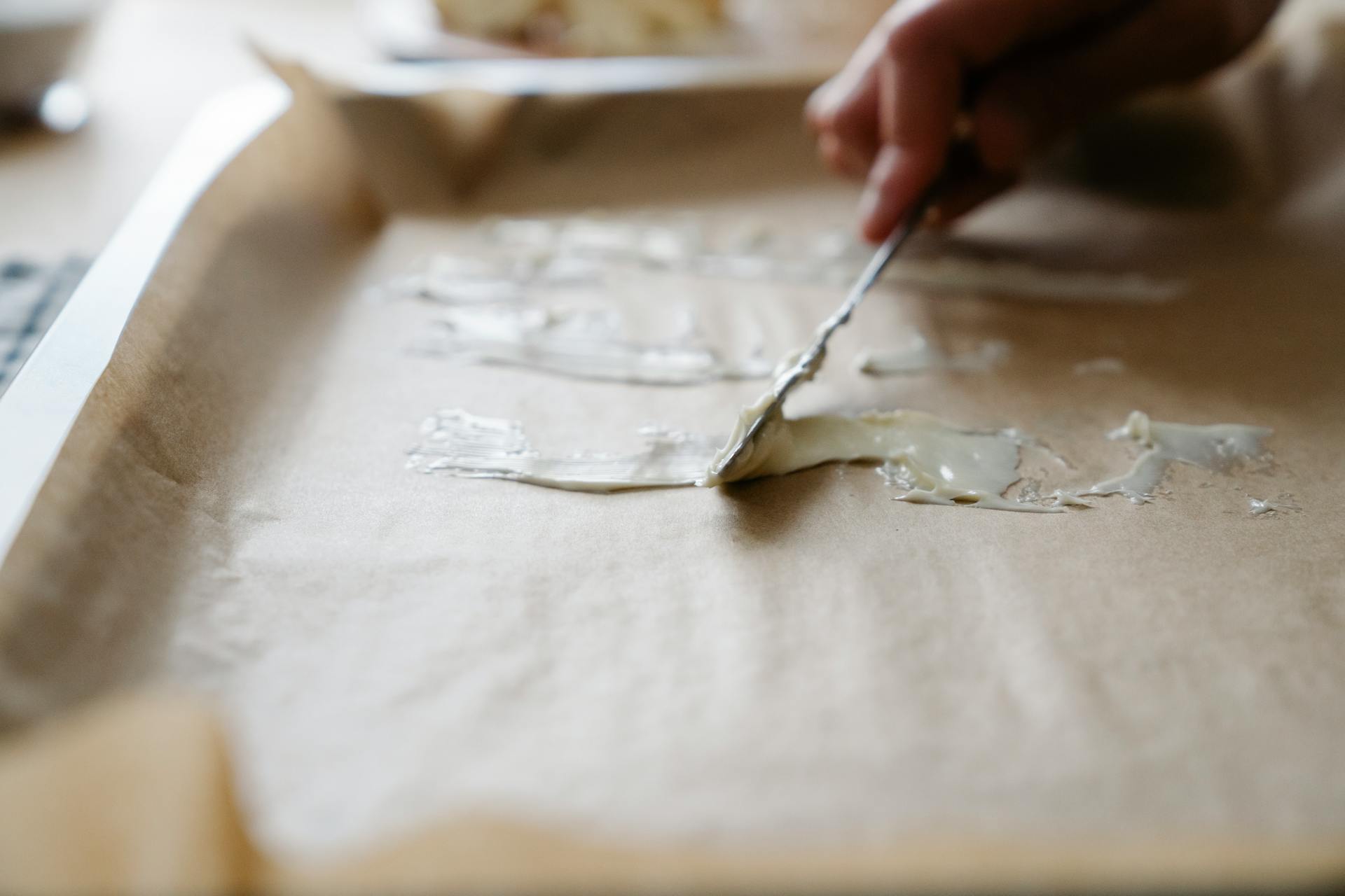 Close-up of hand spreading butter on baking tray lined with parchment paper, preparing for baking.
