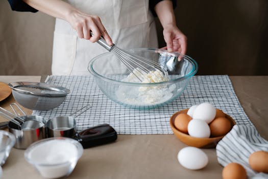 Hands whisking ingredients in a glass bowl, preparing a baking recipe with eggs and flour.