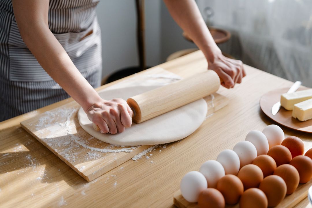 Home Bakery Equipment: What You Actually Need to Start (Under ₹15,000) 4 A woman rolling dough on a floured wooden board in a home kitchen, with baking trays and tools visible in the background — representing a home baker preparing products for sale.