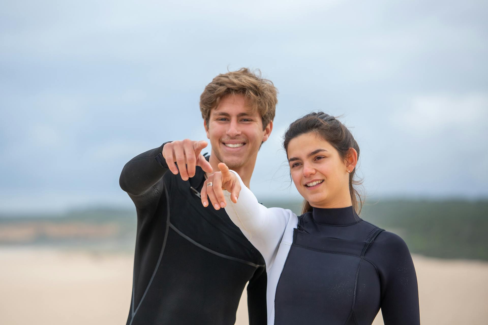 Young couple in wetsuits smiling and pointing on a beach in Portugal, conveying joy and adventure.