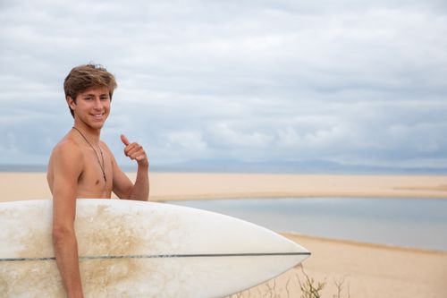 Free Smiling young surfer with a surfboard on a sandy beach in Portugal, giving a thumbs up. Stock Photo