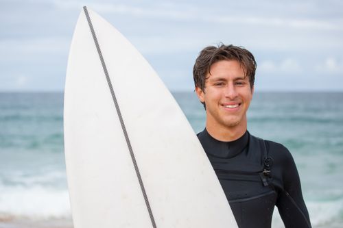 Free A smiling surfer in a wetsuit holding a surfboard by the ocean on a clear day in Portugal. Stock Photo