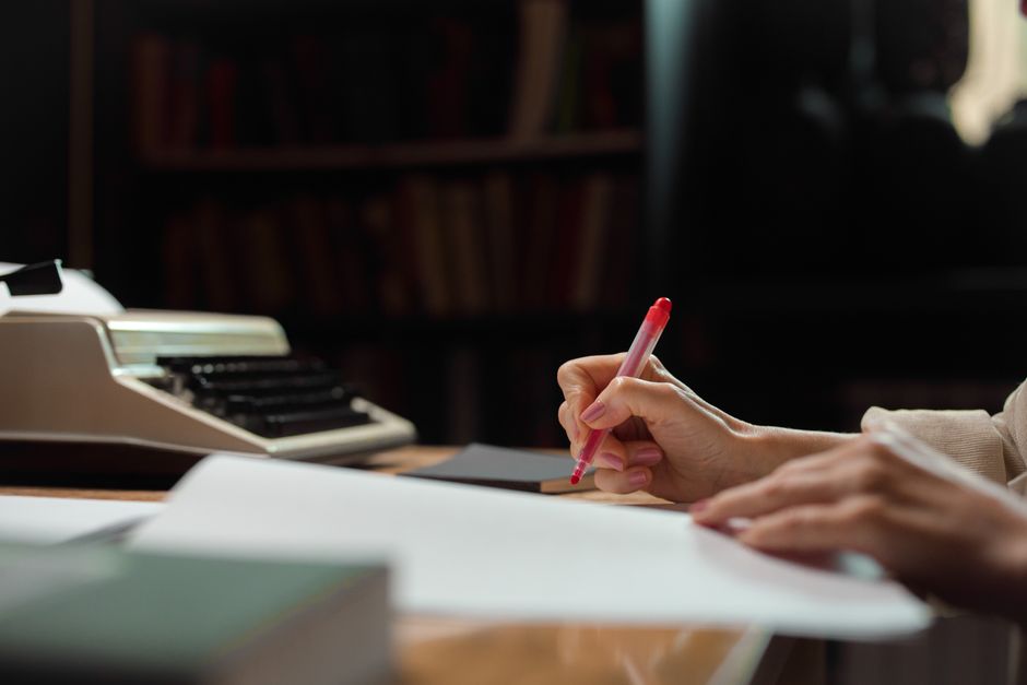 A hand writing with a pen in a cozy library setting with a vintage typewriter.
