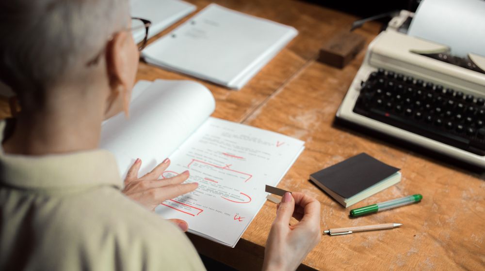 An adult woman reviewing a script with red pen marks at a wooden desk with a typewriter.