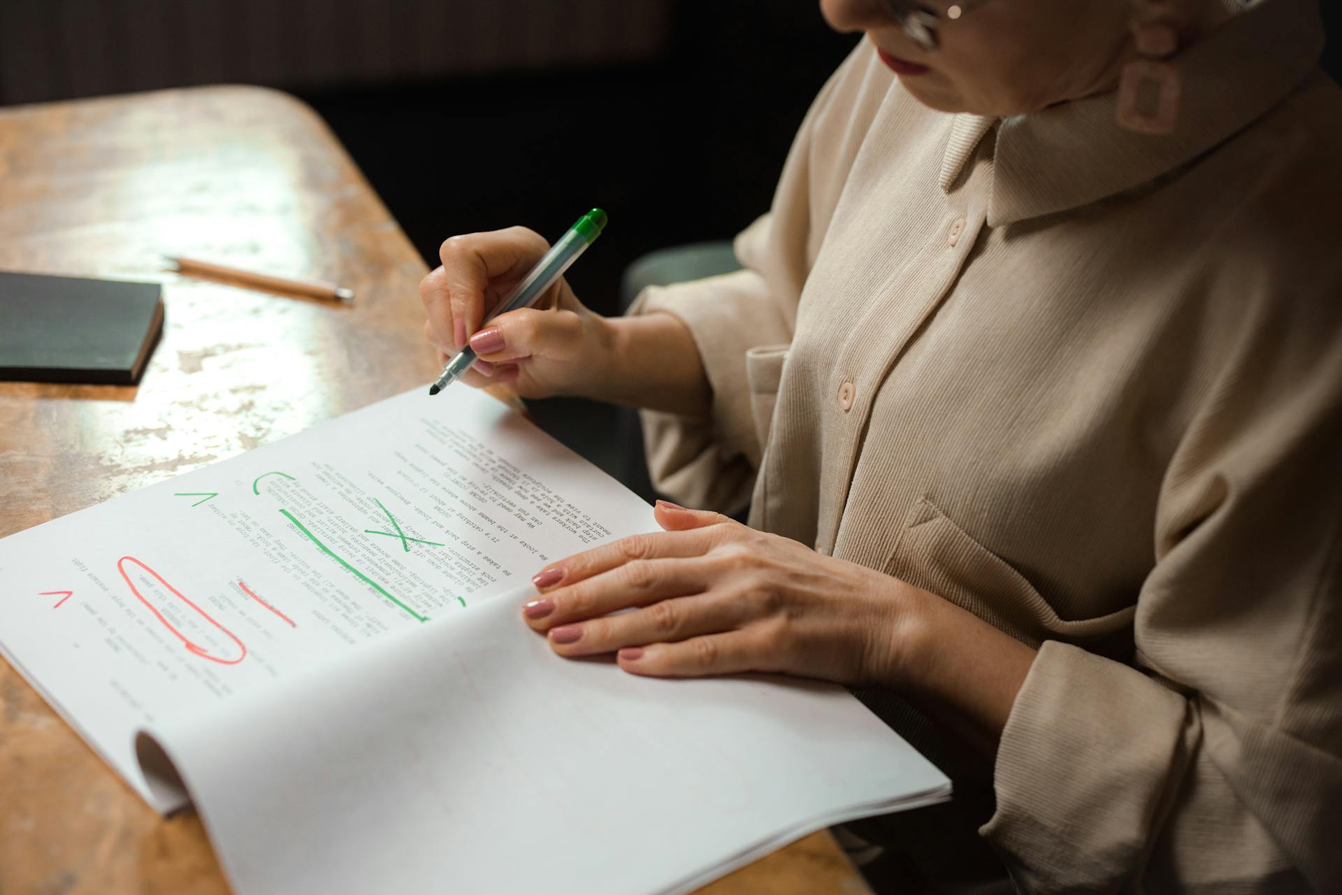 Close-up of a senior woman editing a manuscript at a desk with a green marker indoors.