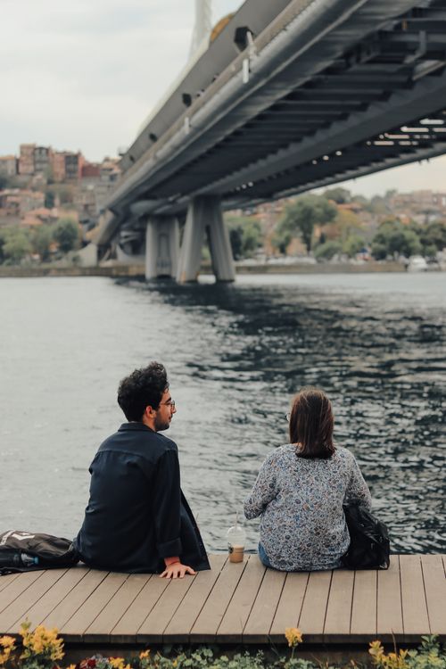 Free A couple enjoying a peaceful moment on a wooden dock by the river with a bridge overhead. Stock Photo