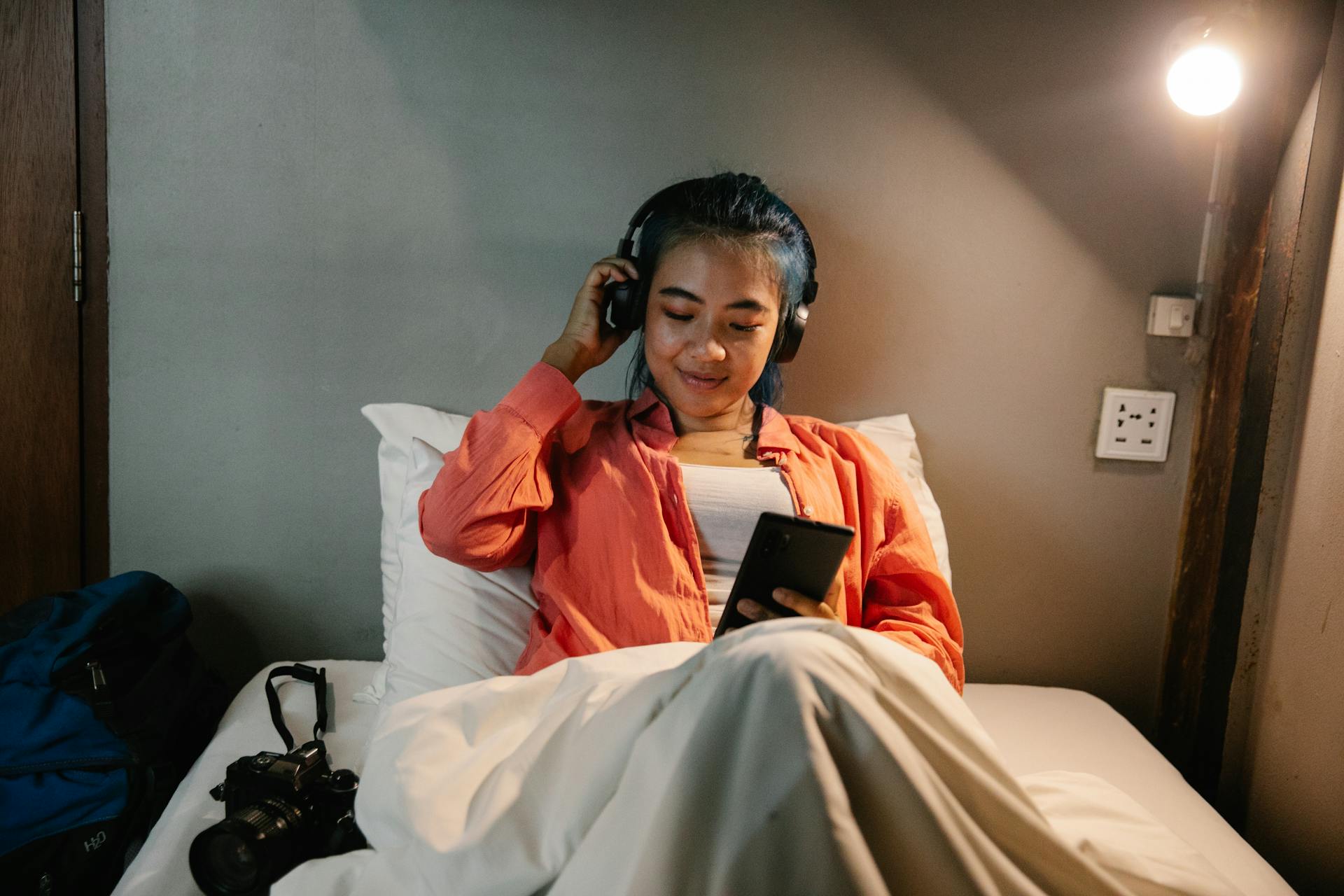 Smiling ethnic woman using smartphone and headphones lying on bed