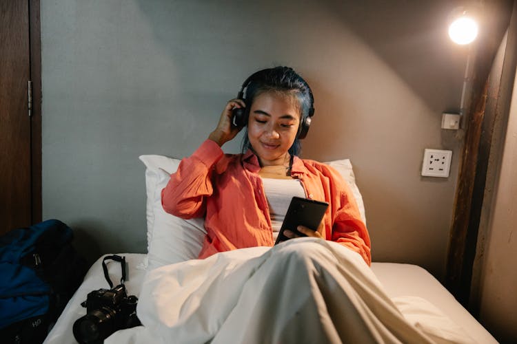 Smiling Ethnic Woman Using Smartphone And Headphones Lying On Bed