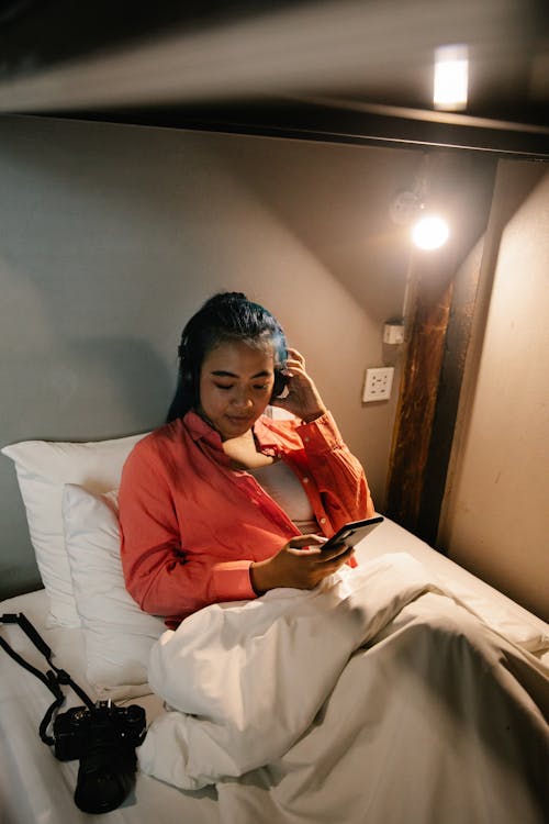Calm young ethnic female resting in comfortable bed under blanket with camera and browsing mobile phone while listening to online music in earphones