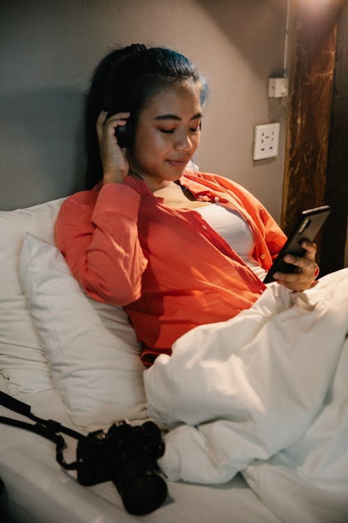 Young ethnic content female resting in comfortable bed under white blanket with camera and surfing mobile phone while listening to music in earphones