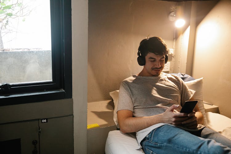 Smiling Man Resting On Bed And Using Smartphone And Headphones