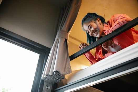Low angle of young ethnic cheerful female resting on bunk bed and looking down from top tier of bed near window with curtain