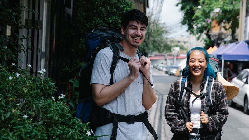 Two smiling young backpackers walking down a leafy street with their travel packs
