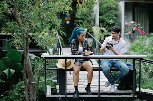 Young male photograph in casual clothes adjusting camera and talking with girlfriend model with blue hair and in checked jacket sitting on wooden bench in suspension footbridge in green garden