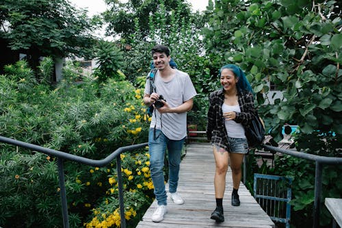 Free Full body of happy multiracial young friends with photo camera and wearing casual clothes walking on wooden footbridge in blooming park Stock Photo