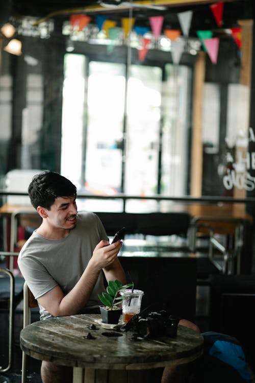 Young positive male in casual clothes messaging with friends on smartphone while sitting at table in cafe