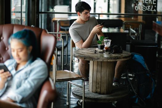 Concentrated male text messaging on cellphone while sitting at table with beverage and photo camera in cafeteria with blurred Asian woman