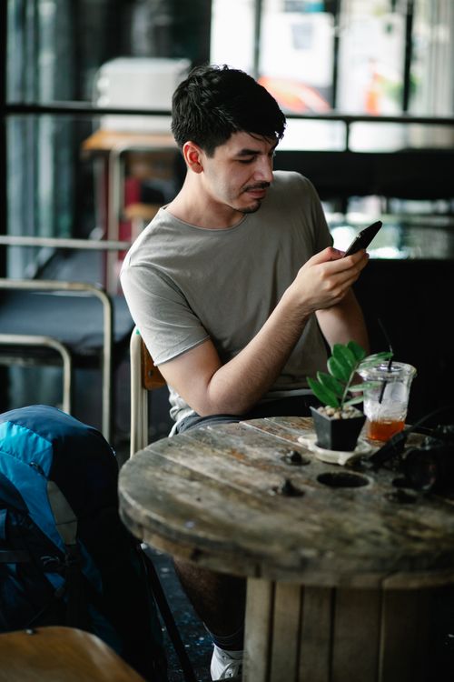 Free Thoughtful male text messaging on cellphone while sitting at round wooden table with refreshing beverage and potted plant in cafeteria Stock Photo