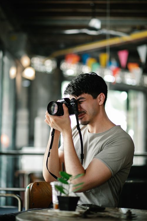 Free Focused male photographer taking picture with modern photo camera while sitting at table with potted plant in cafeteria on blurred background Stock Photo