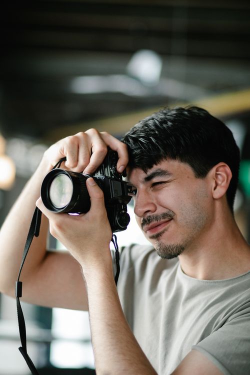 Free Side view of focused male photographer in casual clothes taking picture on professional photo camera in cafe on blurred background Stock Photo