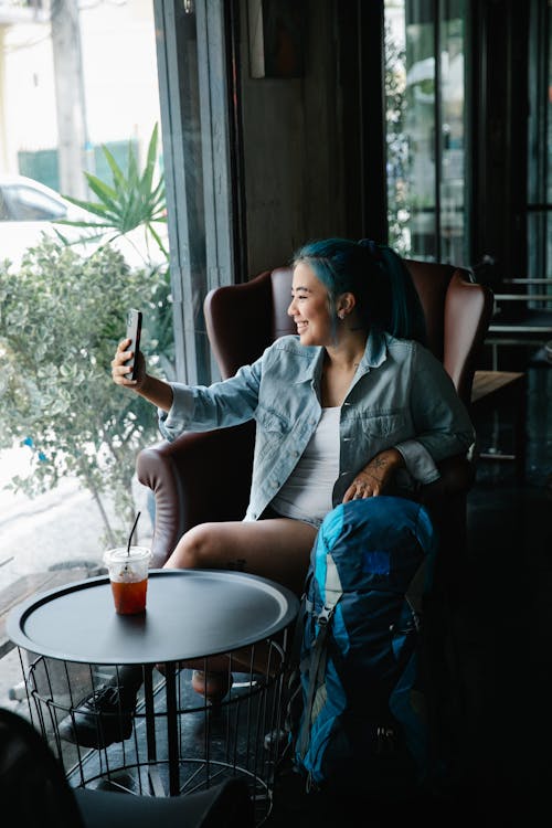 Free Cheerful Asian female in stylish outfit taking self portrait on smartphone while sitting in armchair in table with beverage in cafeteria Stock Photo