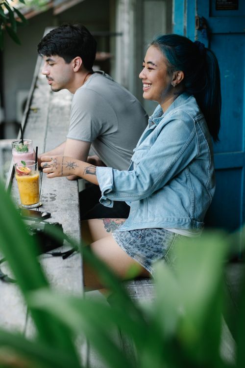 Free Side view of cheerful multiracial couple sitting at wooden table with refreshing drinks on veranda of cafeteria with blurred green plants Stock Photo