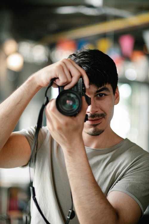 Focused unshaven male photographer taking photo with modern photo camera while looking at camera in light cafe on blurred background