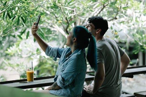Side view of content multiethnic couple taking self portrait on smartphone while sitting at table with refreshing drink on terrace of cafe near green plants