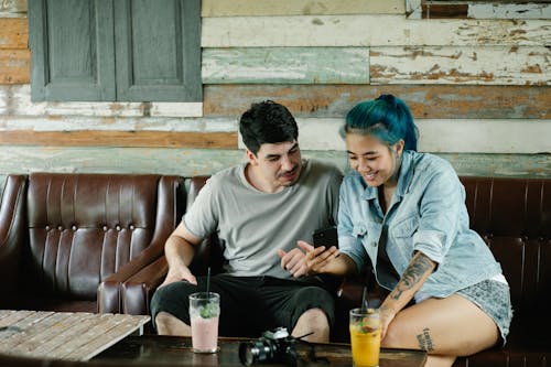 Free Positive multiracial couple surfing modern cellphone while sitting on couch at table with refreshing drinks in cafe with wooden wall Stock Photo