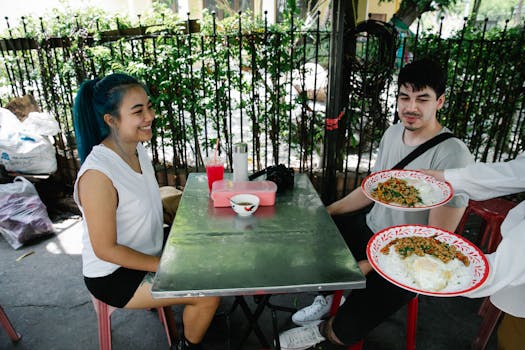 Crop anonymous waiter serving plates of delicious food to positive multiracial couple sitting at table in outdoor cafeteria on summer day