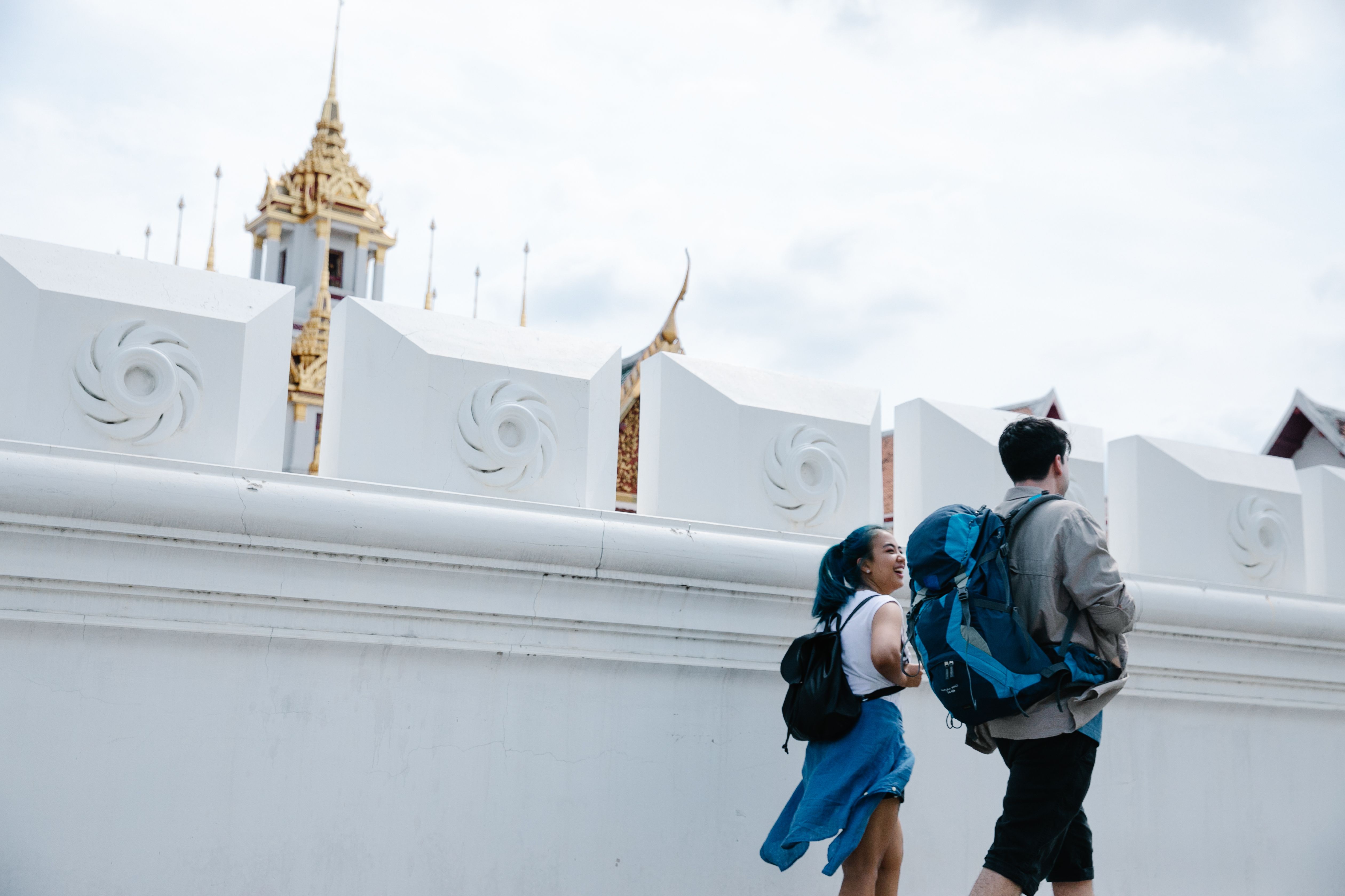 Deux touristes marchant devant un mur blanc à Bangkok avec un temple doré visible.