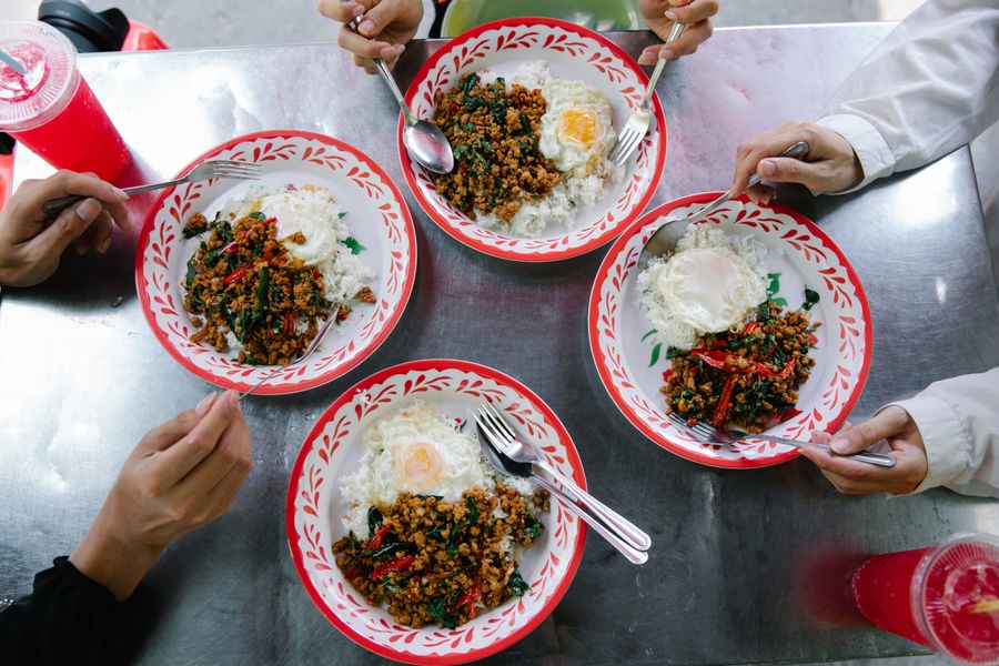 Overhead view of four plates of Thai basil chicken and rice shared at a street food table.